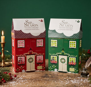 Two Christmas-themed house-shaped gift boxes labeled 'Sip the Season' on a rustic wooden surface, surrounded by pine branches, berries, a lit candle, and a feather quill against a dark green background.